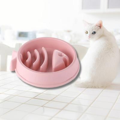 Il y a une gamelle rose avec forme de poisson à l'intérieur. Il y a un chat blanc avec un œil vert et un bleu qui est assis de dos sur une table carrelée blanche et qui tourne juste la tête face caméra.